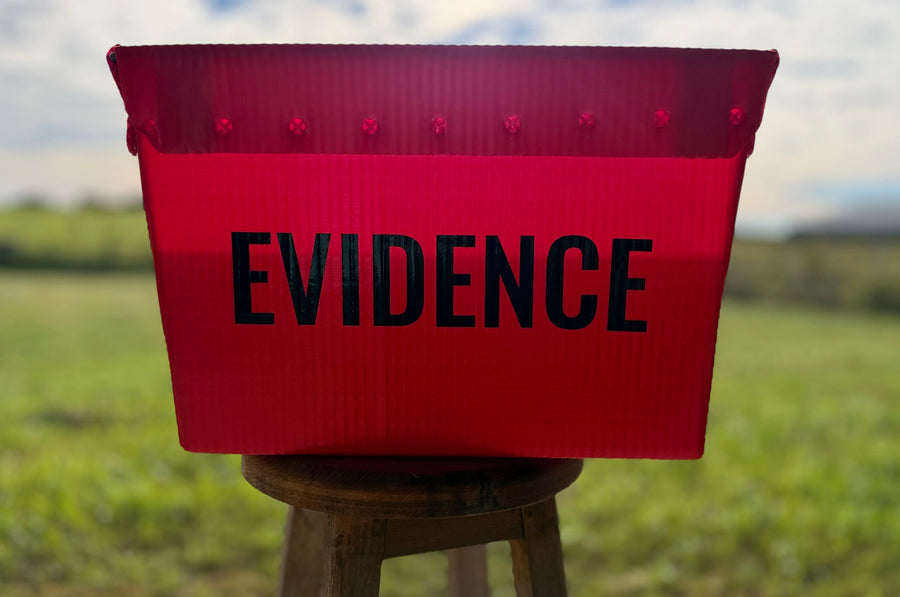 A red corrugated plastic evidence bin with the word 'EVIDENCE' printed in black capital letters, sitting on a wooden stool in an outdoor grassy field.