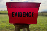 A red corrugated plastic evidence bin with the word 'EVIDENCE' printed in black capital letters, sitting on a wooden stool in an outdoor grassy field.
